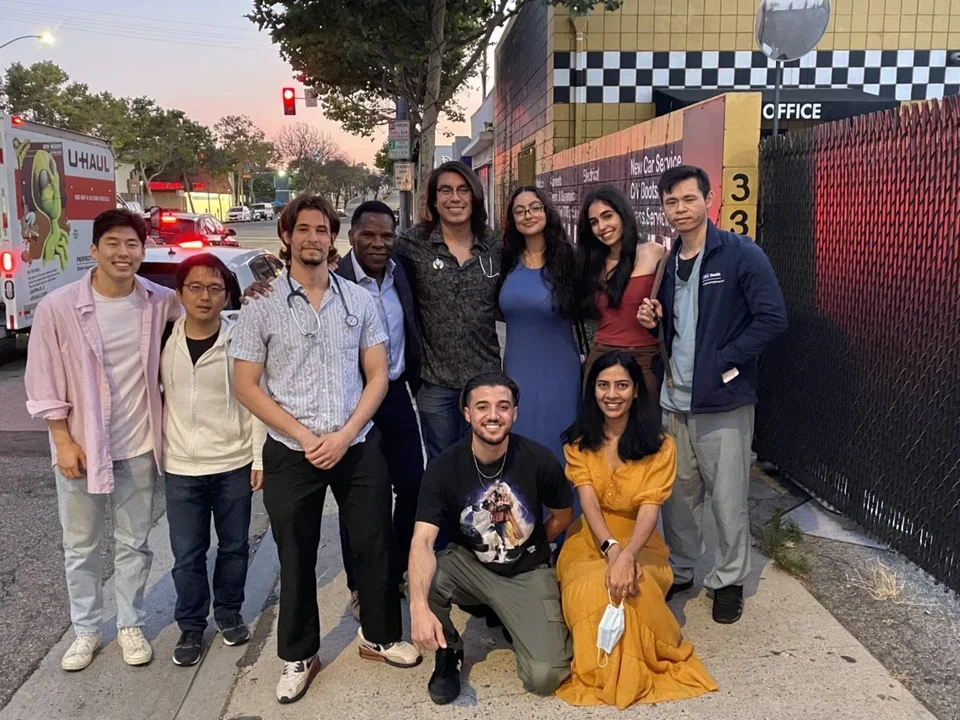 Ajijola Lab members posing together on a sidewalk at sunset in front of a mural-covered wall.